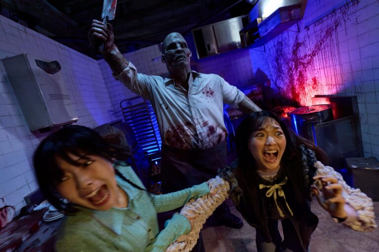 Two screaming guests flee a blood-spattered zombie butcher raising a cleaver in a tiled kitchen