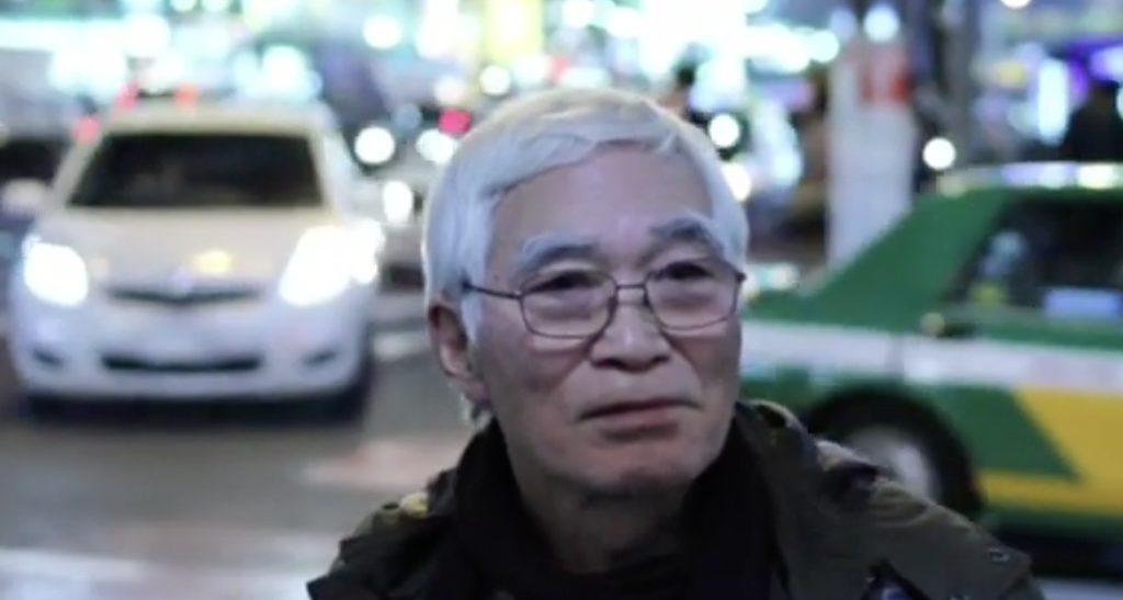 Adachi Masao, old, with white hair, stands in front of a blurry background showing cars in Shibuya.