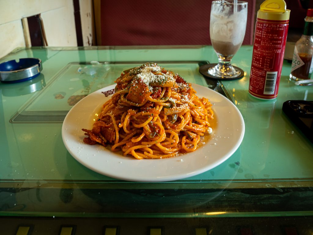 A plate of naporitan topped with grated cheese on a green glass table, beside an ashtray, iced water, and a Tabasco bottle.