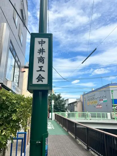 A green signpost reading 中井商工会 on a Nakai street, with a pedestrian bridge and low-rise buildings behind it.