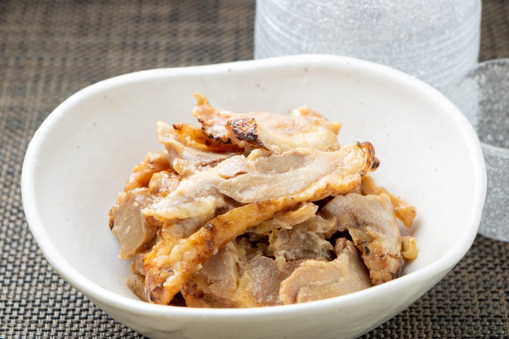 A shallow white bowl of seared chicken slices in ponzu sauce sits on a woven mat beside a frosted glass.