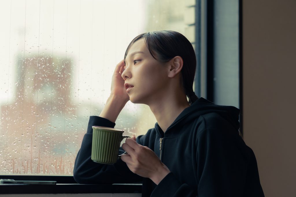 A young woman in a black hoodie holds a mug and looks out a rain-streaked window from a table indoors.