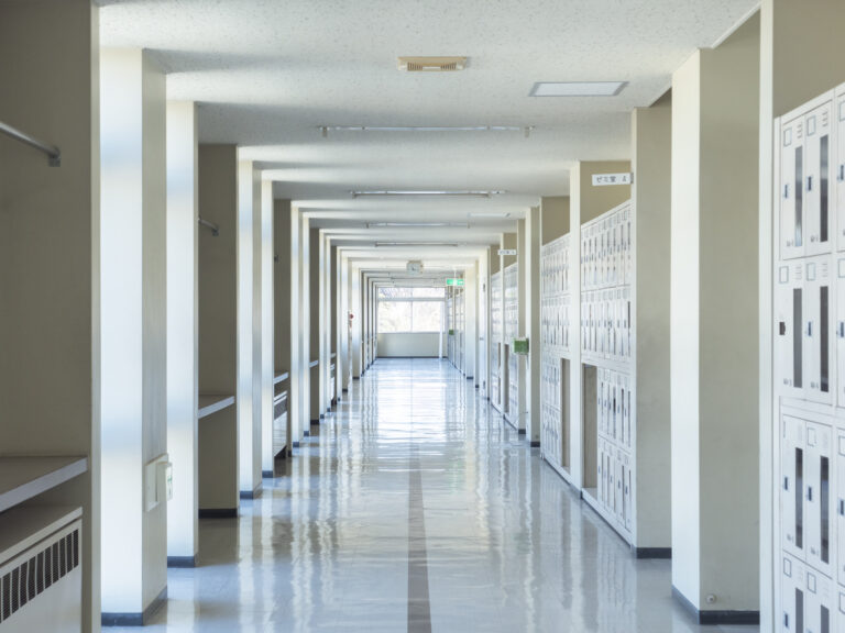 Empty sunlit corridor in a Japanese school, lockers lining one wall and classroom signs overhead