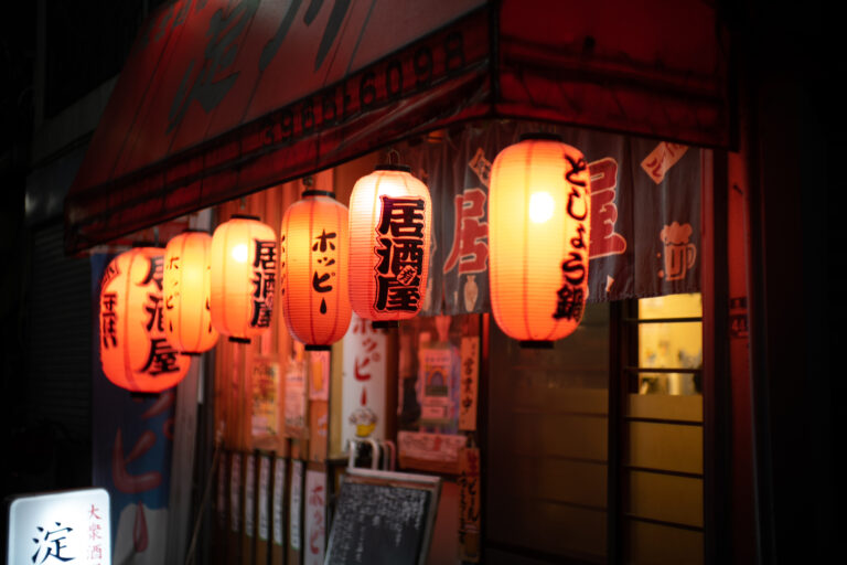 Red paper lanterns reading izakaya and Hoppy glow outside a small Japanese pub at night, above handwritten signs.
