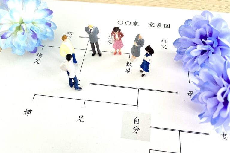 Miniature figures standing on a printed Japanese family tree chart labeled with relatives, beside blue paper flowers