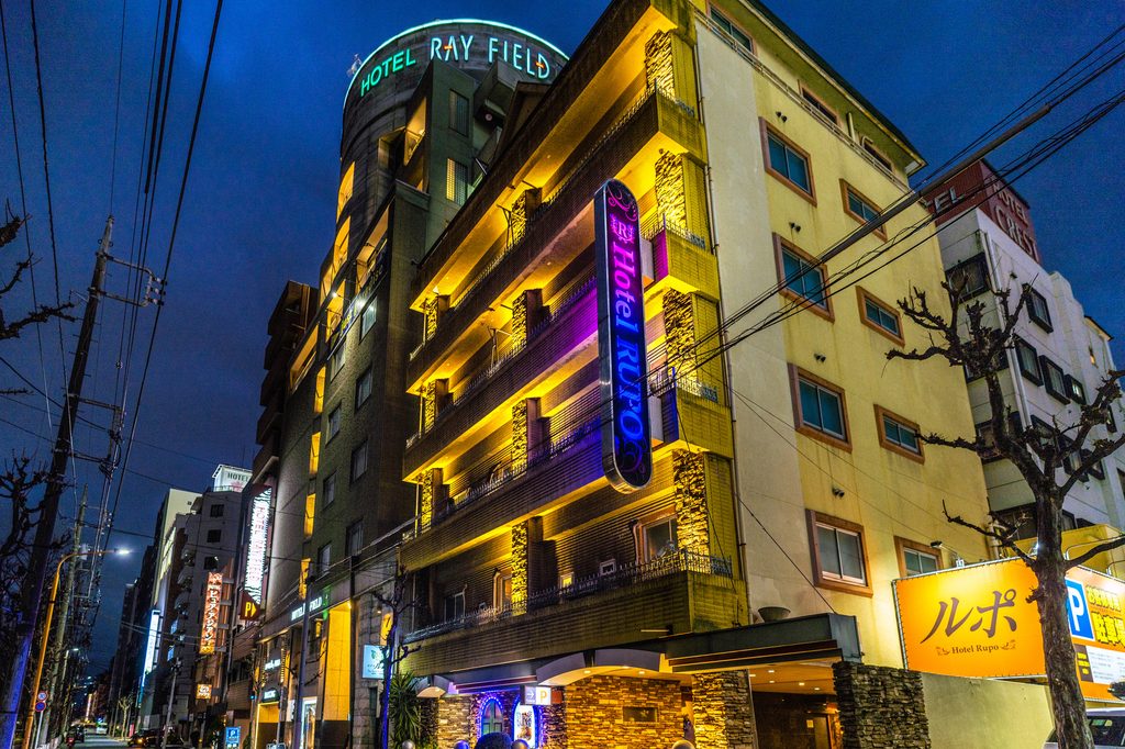 Several love hotels clustered along one street at dusk, their neon signs lit above ground-floor parking entrances.