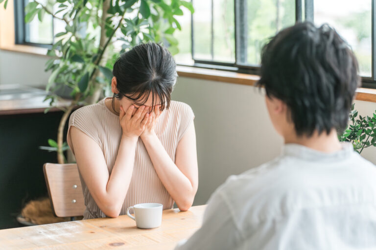 A woman covers her face with both hands at a café table while a man sits across from her