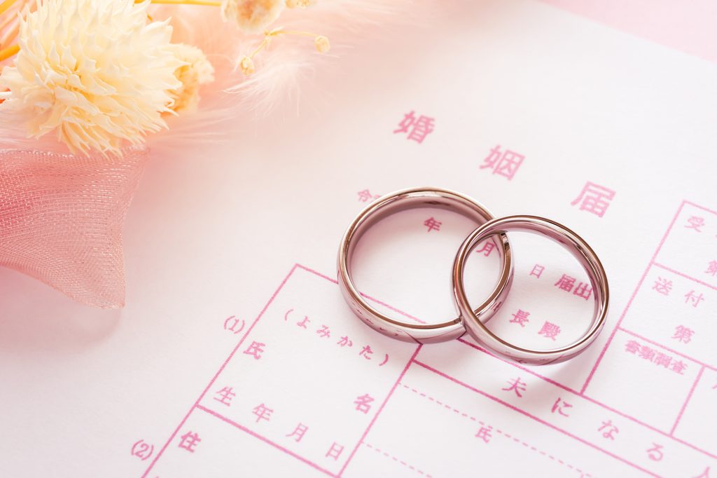 Two wedding bands rest on a pink Japanese marriage registration form next to dried flowers.