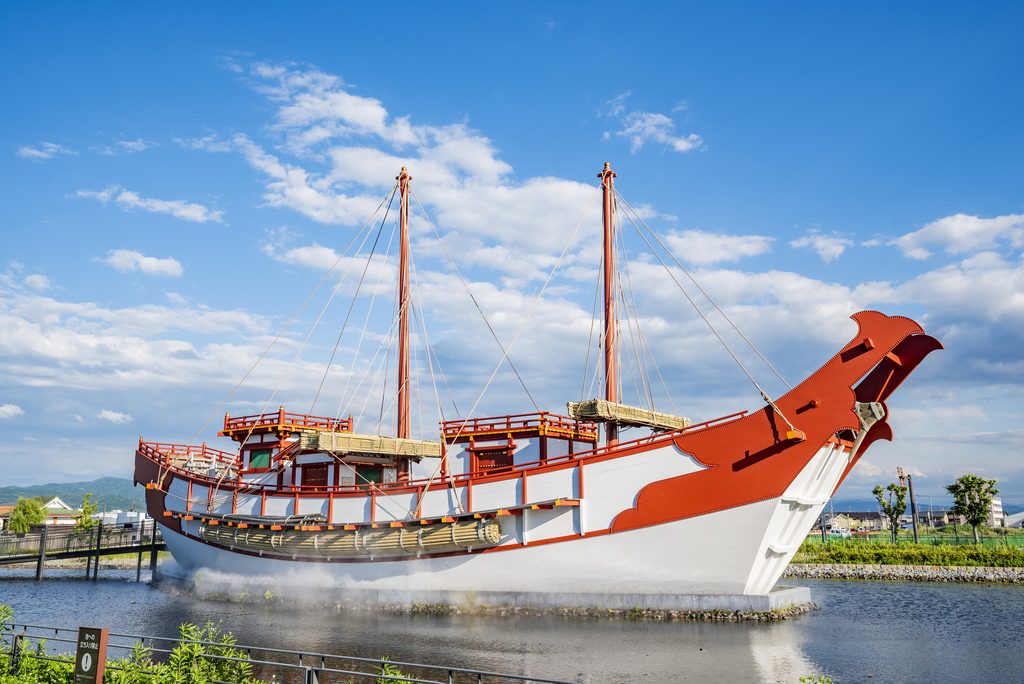A red and white replica of a Tang-era envoy ship with two masts, moored in a pond under a blue sky.
