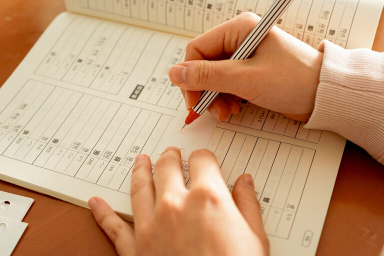 A learner practices kanji in an open Japanese workbook, holding a striped red pen over a numbered practice grid.
