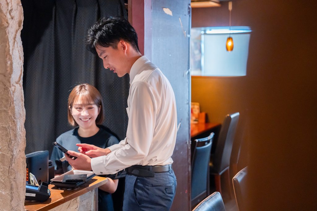 A staff member holds a payment tablet at an izakaya counter with a card reader and receipt printer as a customer waits.