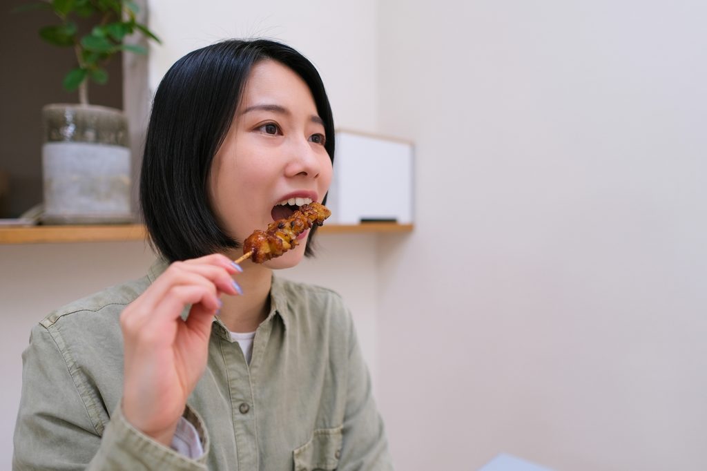 A young woman with a black bob bites grilled chicken straight off the skewer.