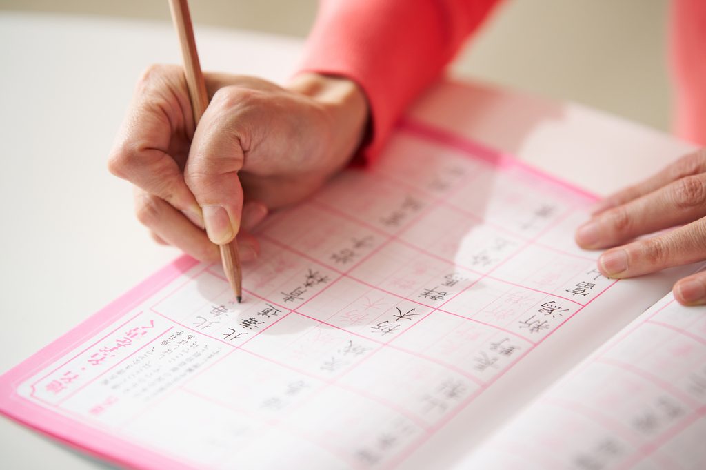 A hand writing kanji in pencil in a pink-bordered kanji practice workbook