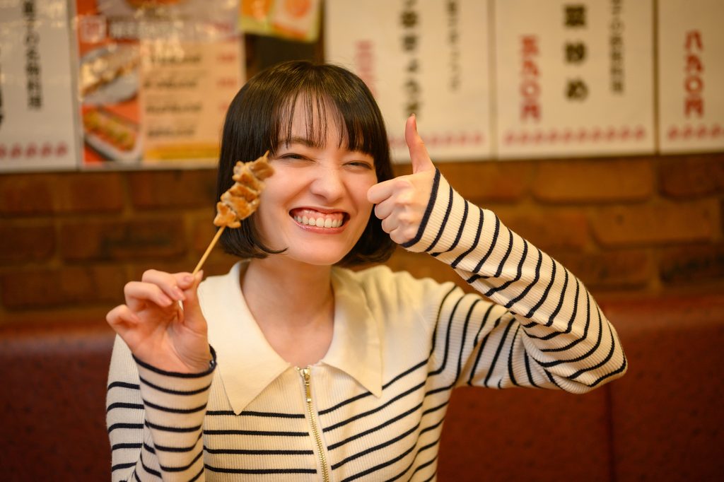 A smiling woman in a striped cardigan holds up a yakitori skewer and gives a thumbs-up in front of an izakaya’s handwritten menu.