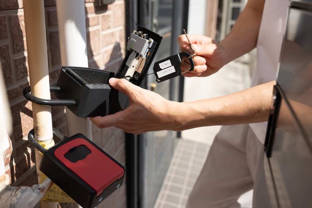 Hands opening a wall-mounted key lockbox beside an apartment entrance, with a second red lockbox below it.
