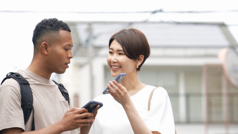 A traveler with a backpack and a Japanese woman stand talking on a residential street, each holding up a smartphone.