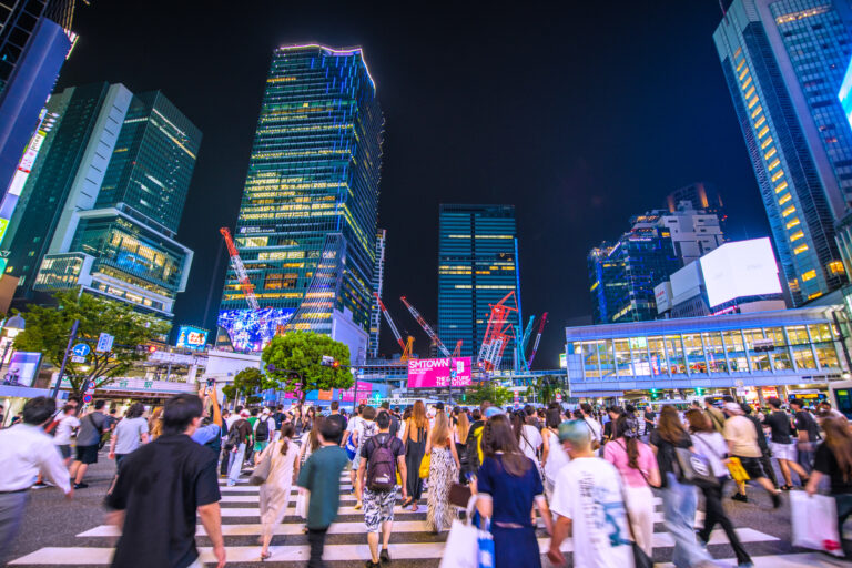 Crowds cross Tokyo's Shibuya Scramble Crossing at night, surrounded by lit high-rises, construction cranes and neon billboards.