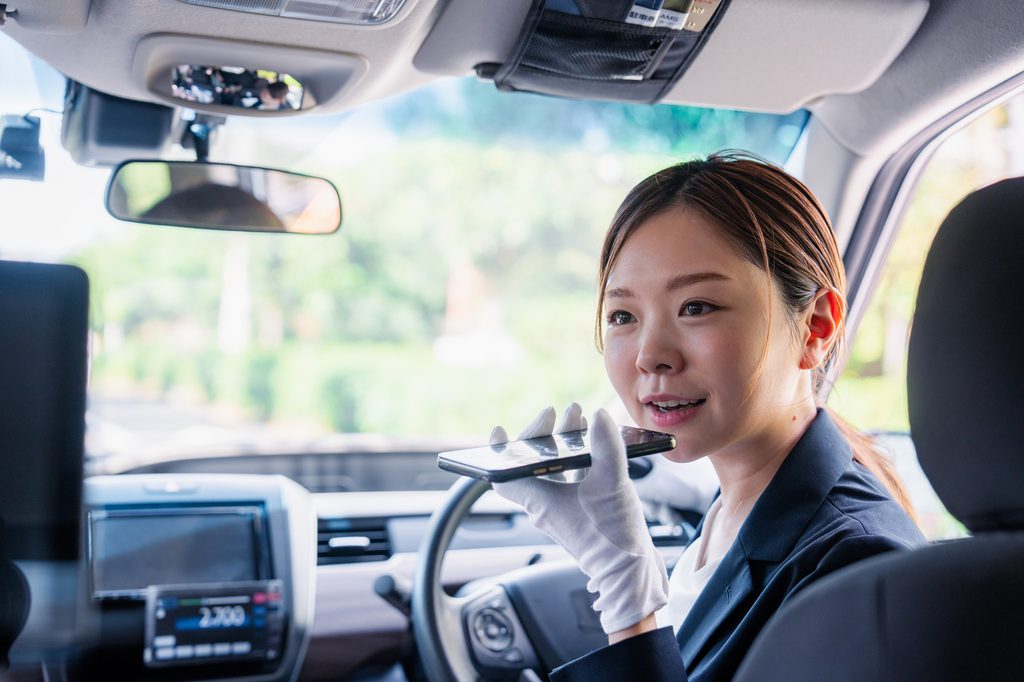 A taxi driver in uniform and white gloves speaks into a smartphone held near her mouth while seated at the wheel.