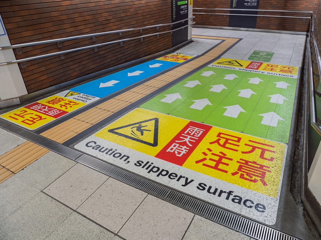 Colored floor decals in a Japanese station reading "Caution, slippery surface" in English and Japanese, with directional arrows.