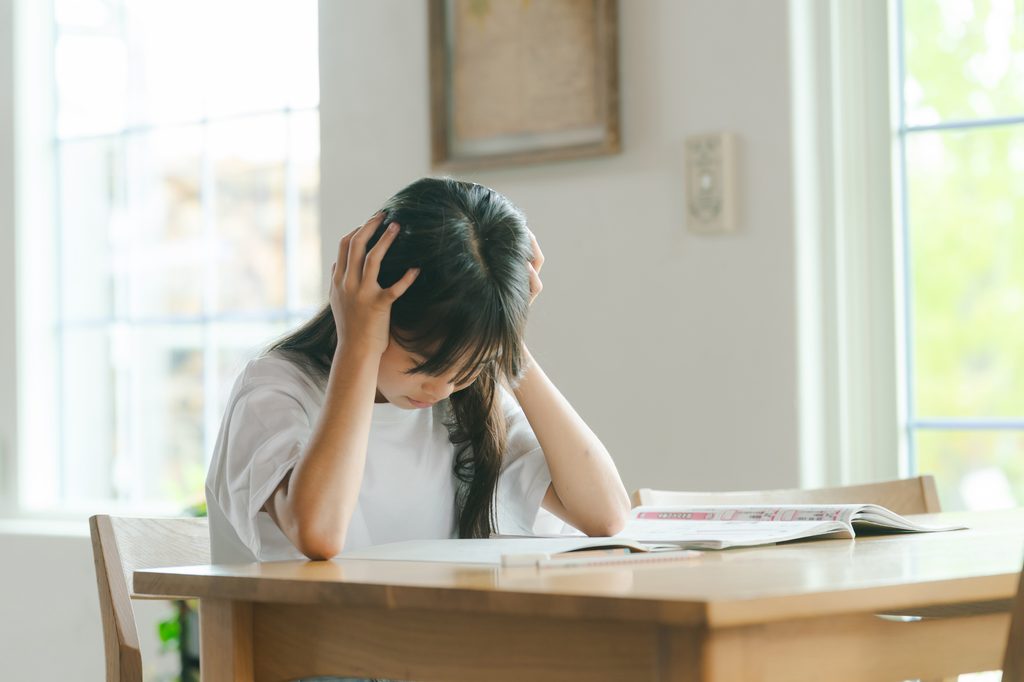 A girl holds her head in frustration over an open workbook at a wooden table