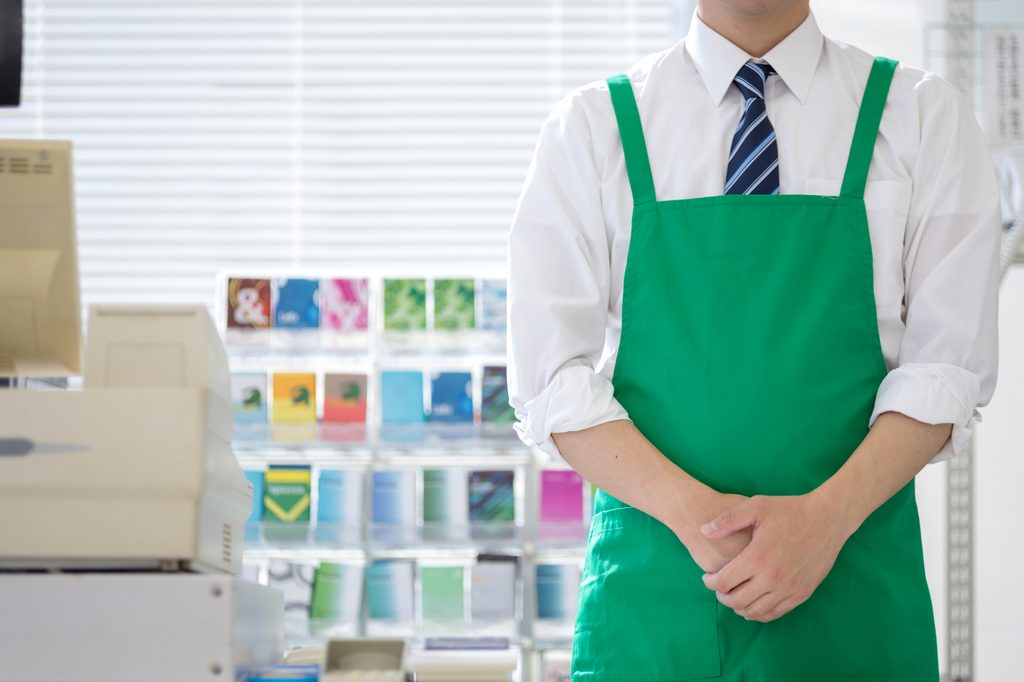 A convenience store clerk in a green apron and tie standing by the register