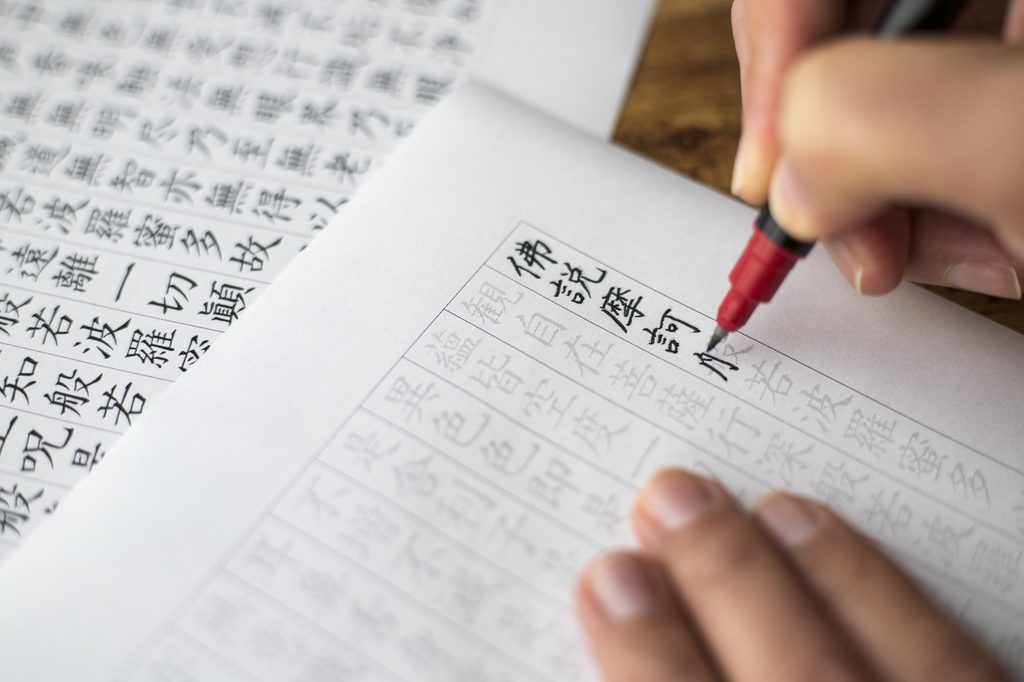 A hand tracing printed kanji of the Heart Sutra in red pen on a sutra-copying sheet.