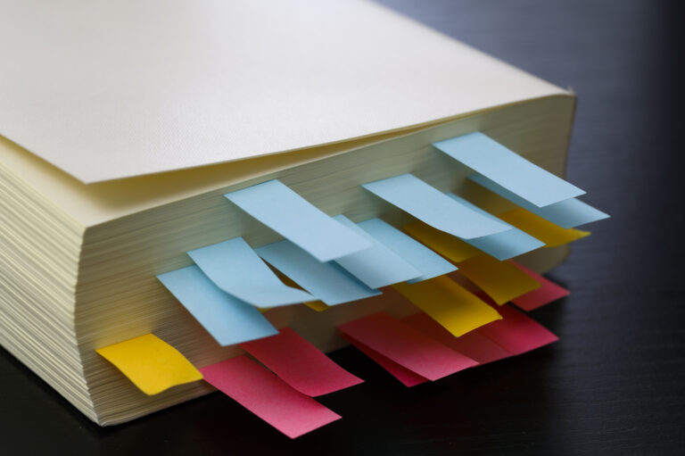 A thick book bristling with blue, yellow and pink page tabs on a dark desk.