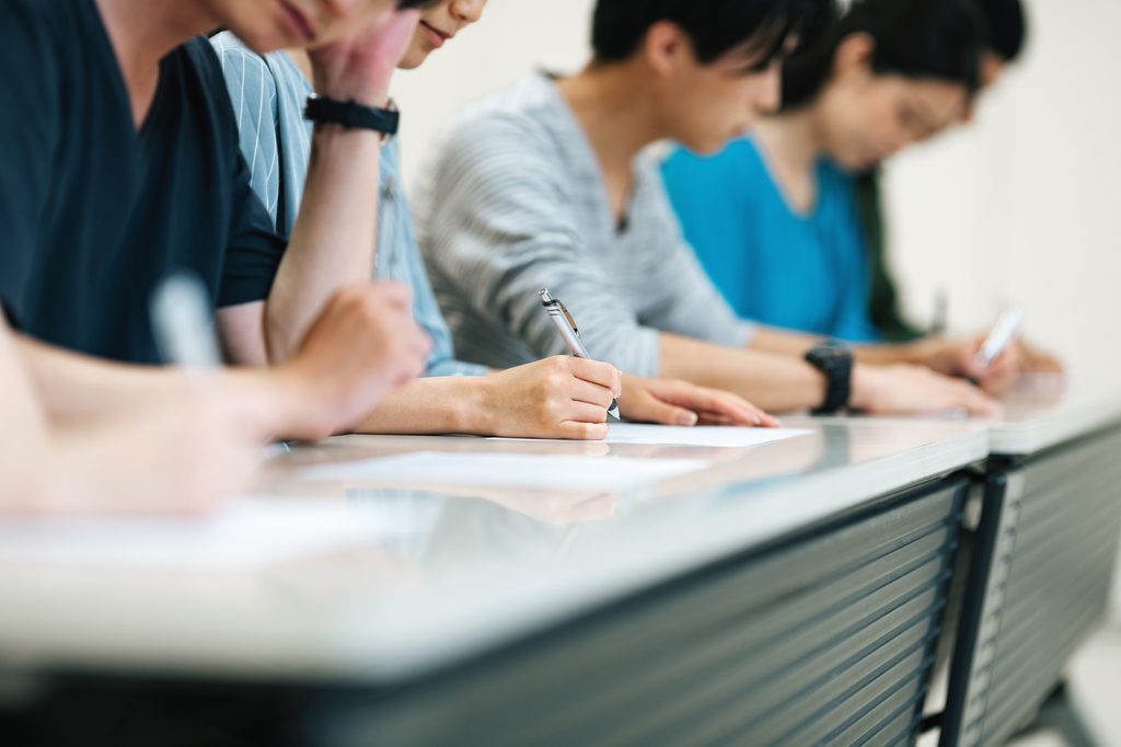 Test-takers sitting in a row, writing answers on paper at a long exam-hall desk