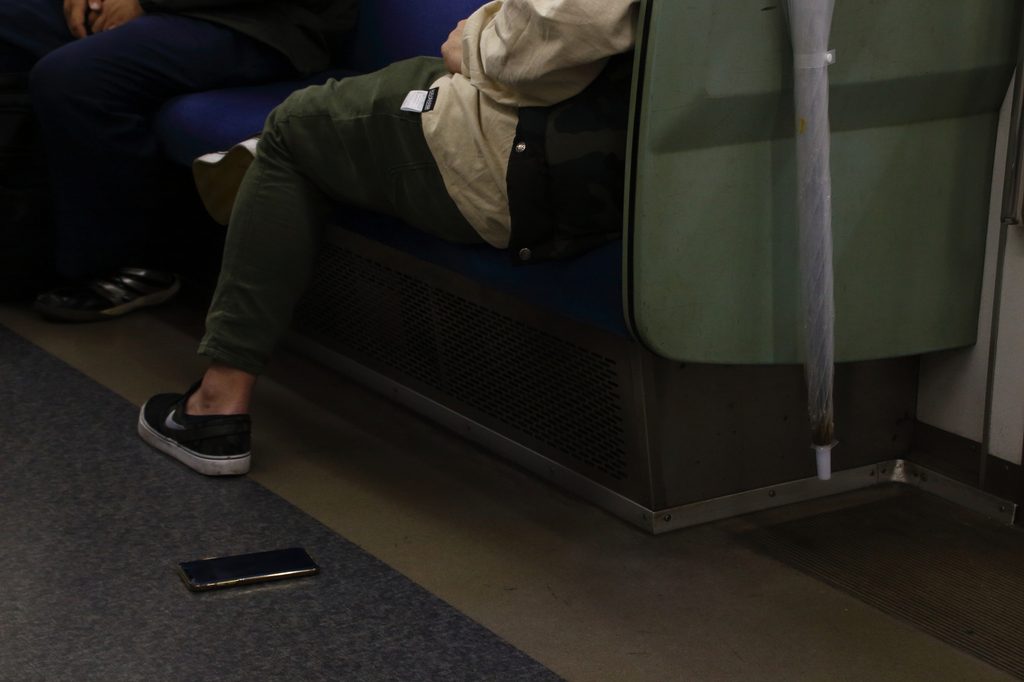 A smartphone lying face-up on the floor of a Japanese train carriage, beside a seated passenger's feet.