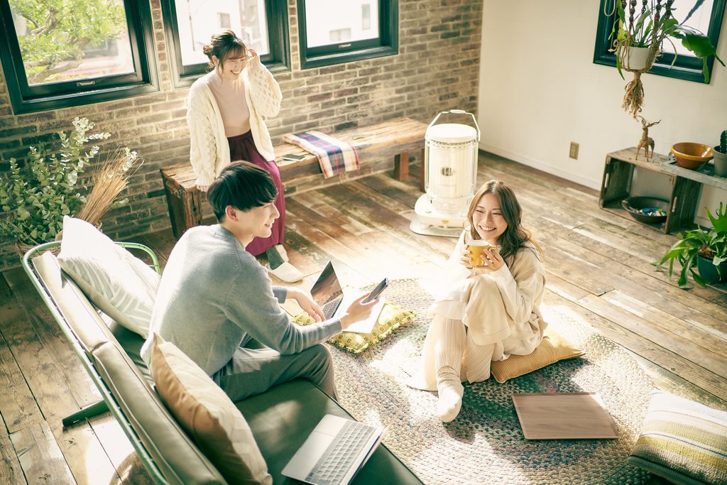 Three young adults share a sunlit, wood-floored room, one at a laptop, one holding a mug, one standing by the window.