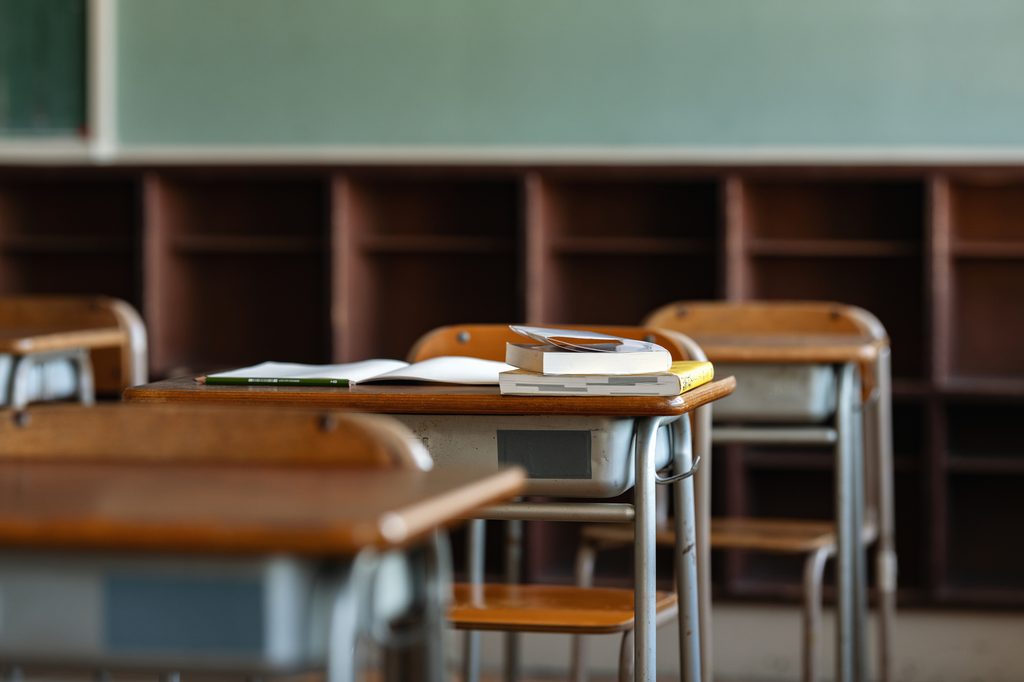 Empty wooden desks in a Japanese classroom, an open notebook and textbooks left on one, blackboard behind
