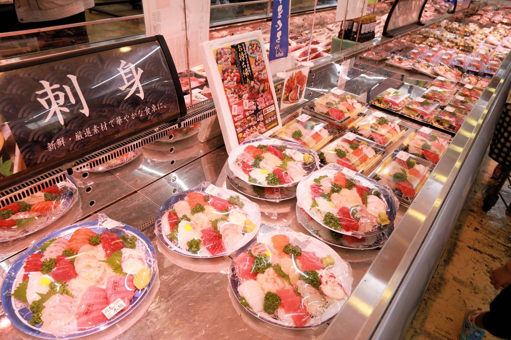Plastic-wrapped platters of assorted sashimi, including salmon and tuna, beneath a "sashimi" sign at a Japanese supermarket