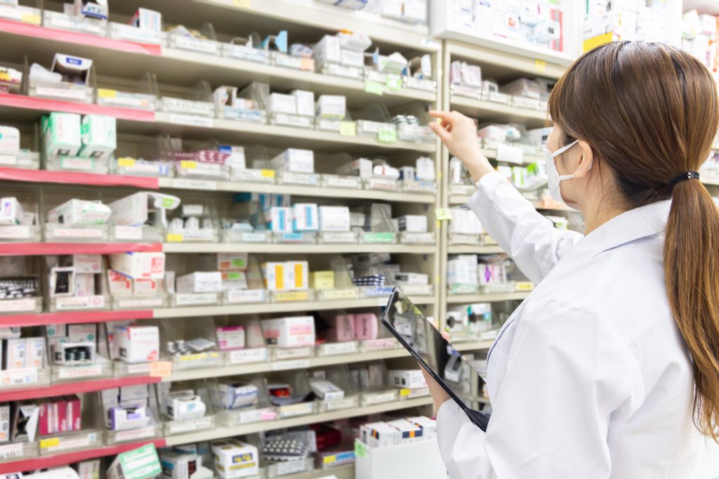 Masked pharmacist in a white coat reaching for medicine boxes on pharmacy shelves
