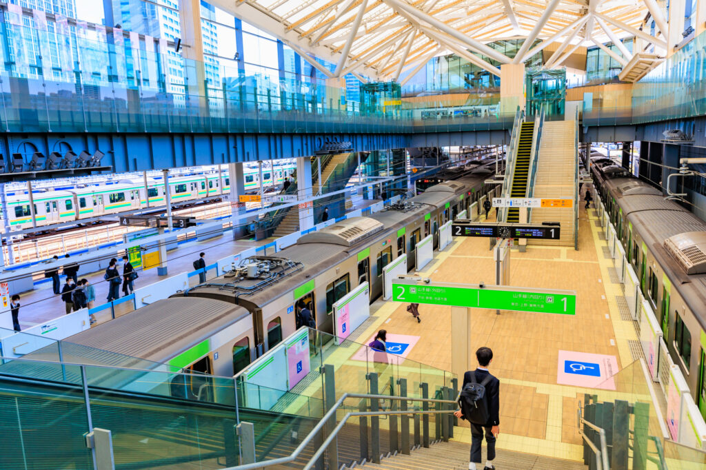 The bright, high-roofed interior of Takanawa Gateway Station, with green-striped Yamanote Line trains at platforms 1 and 2.