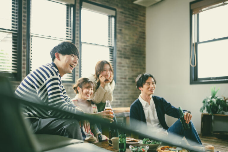 Four young adults laugh together on the floor of a bright brick-walled apartment, drinks and shared plates on a low table.