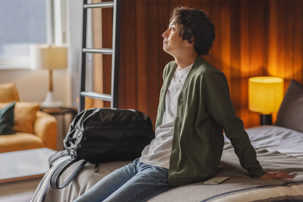 A man sits on the edge of a bed in a small lodging room, backpack beside him and a lamp lit against a wood-panel wall.