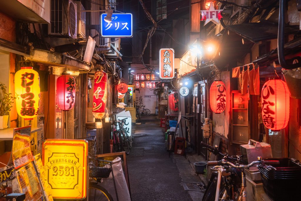A narrow Tokyo alley at night lined with small bars and izakaya under red paper lanterns and signs