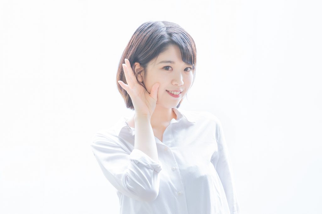A woman in a white shirt smiles and cups a hand behind her ear, as if straining to hear.