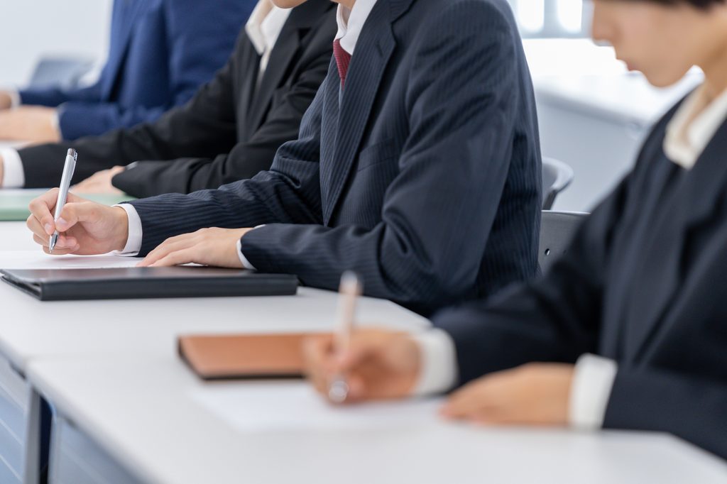 Test-takers in dark suits write with pens at a long row of white desks in an exam room.