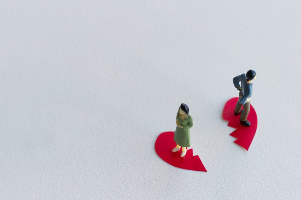 Two small figurines of a man and a woman standing apart, each on one half of a torn red paper heart