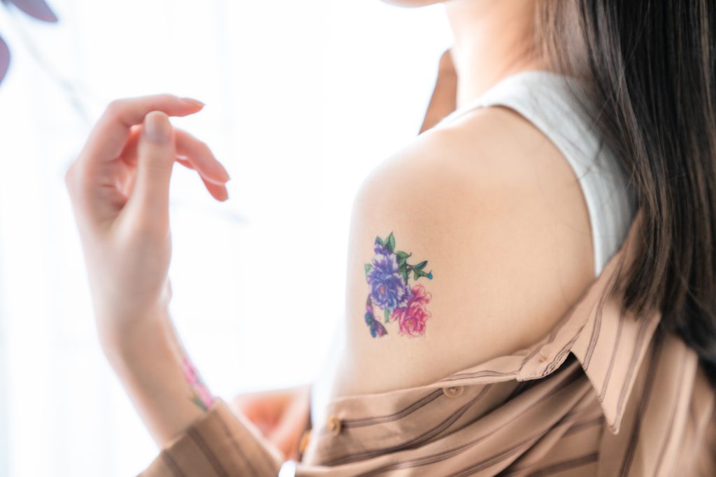 Woman slips a shirt off her shoulder to show a small color tattoo of purple and pink flowers on her upper arm.