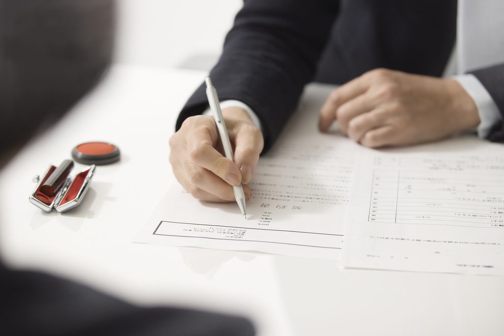 A person in a suit fills out a Japanese contract form in pen, with a hanko seal and red ink pad on the desk beside them.