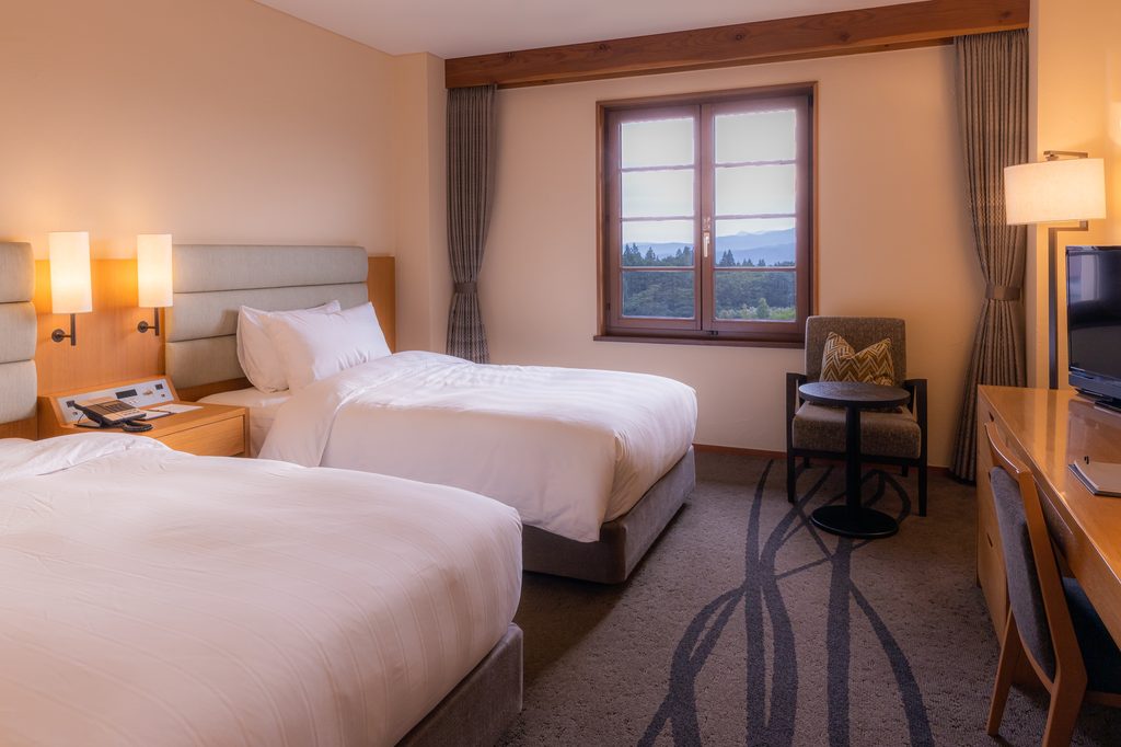 A conventional twin-bed hotel room with a writing desk, armchair and a window looking onto forested hills.