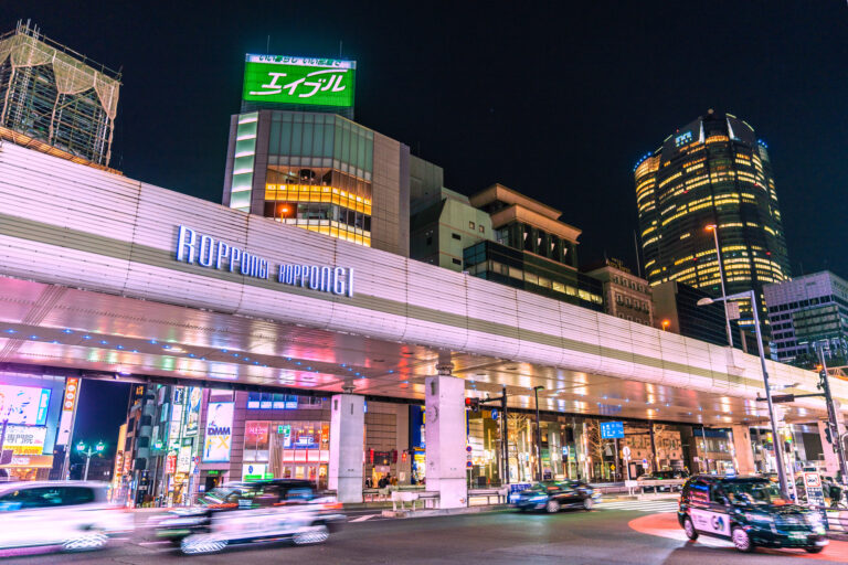 Traffic streaking under the illuminated Roppongi Crossing overpass at night, with neon signs and Roppongi Hills Mori Tower lit behind it.