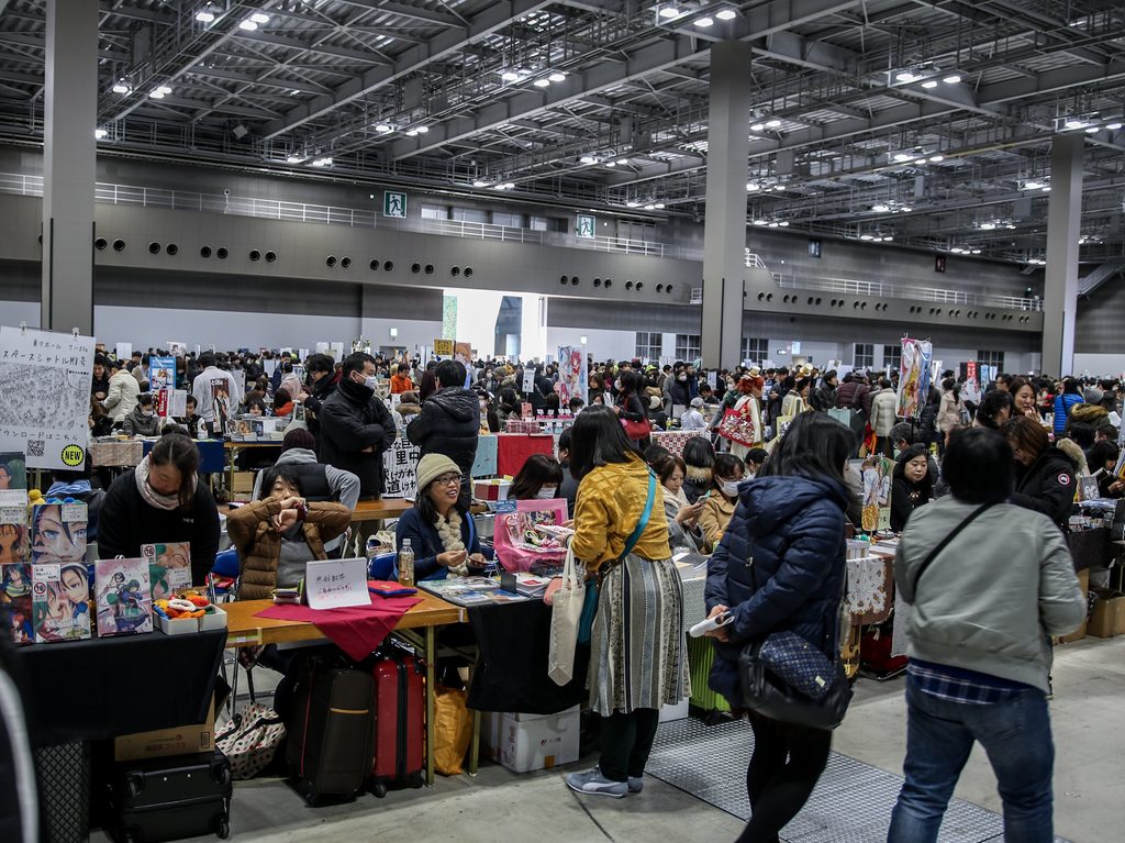 Rows of seller tables and crowds of shoppers filling a large exhibition hall at a dōjinshi convention.