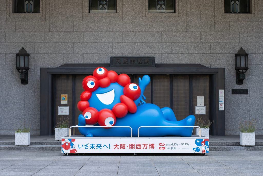 A sculpture of Myaku-Myaku, the Osaka-Kansai Expo 2025 mascot, reclining outside Osaka City Hall beside an Expo banner.