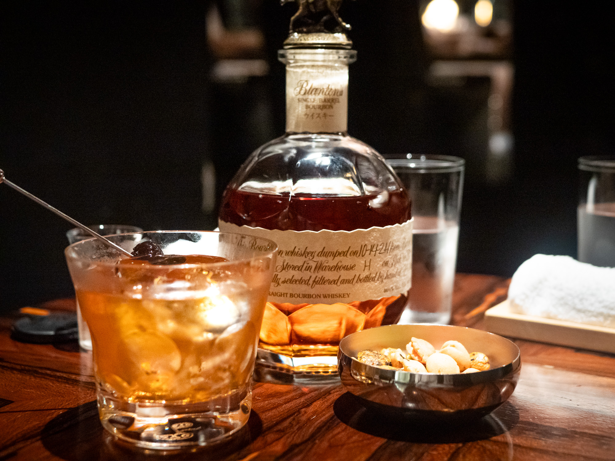An Old Fashioned cocktail on a dark wood bar counter beside a bourbon bottle
