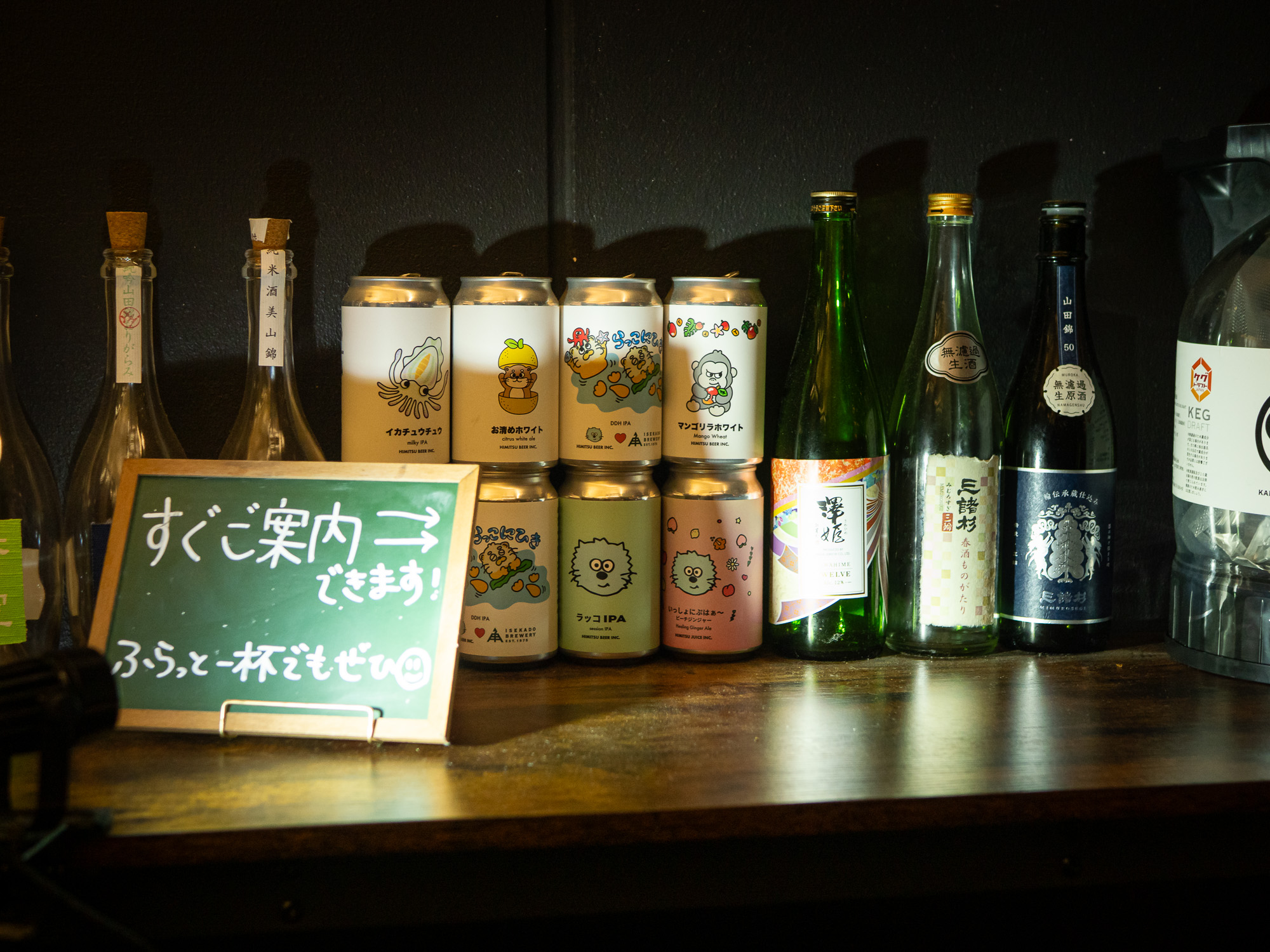 Sake bottles and craft beer cans lined up on a warmly lit counter shelf