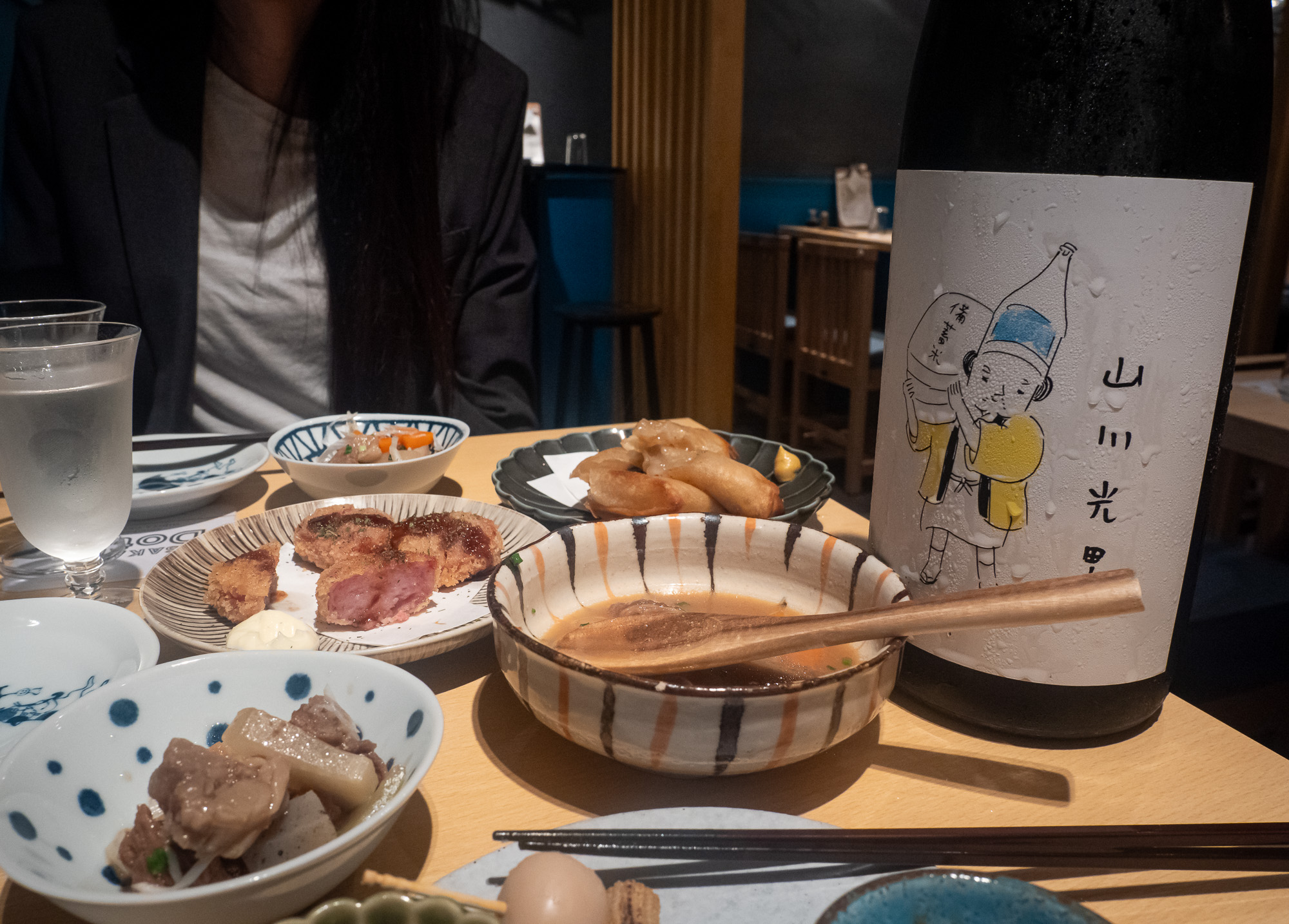 A table of small dishes and a simmering oden pot beside a bottle of sake