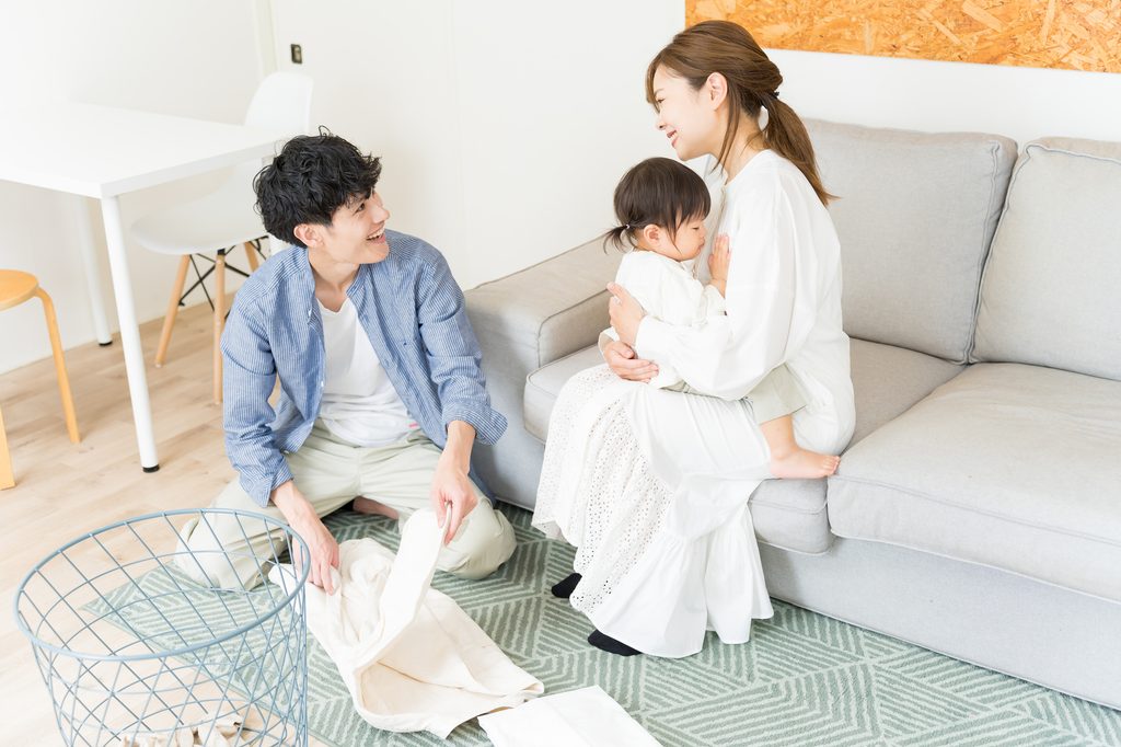 A father folds laundry on the floor while a mother holds a toddler on the sofa, both smiling at each other